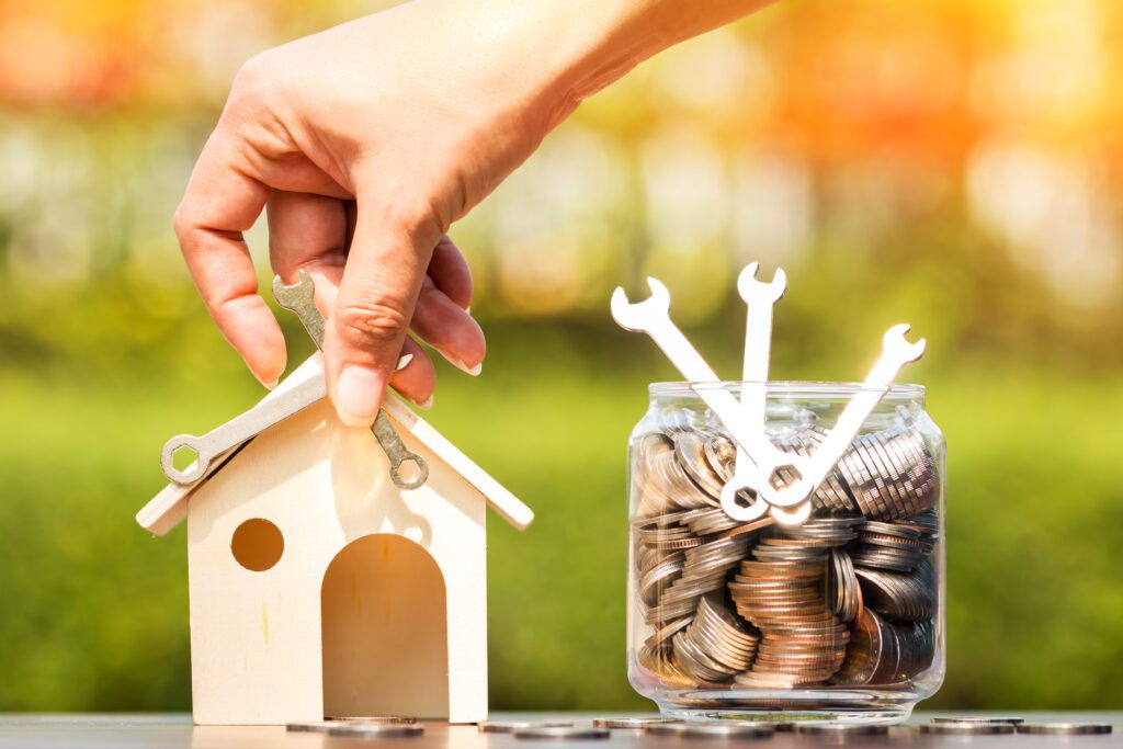 Person holds small wrenches over wooden model house next to coin jar | Home repair financing